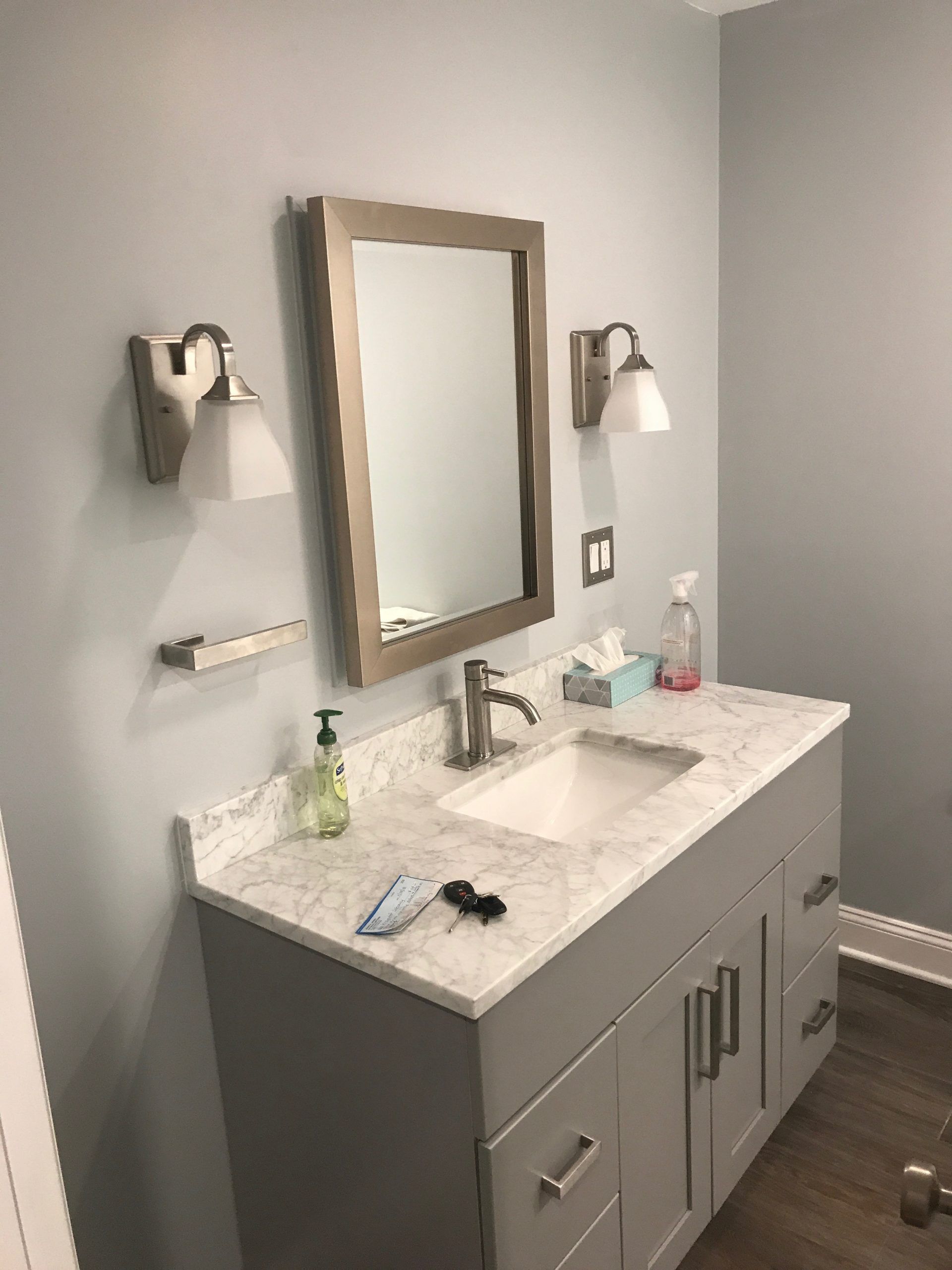 Modern bathroom vanity with double sink, wall mirror, sconces, and gray cabinets with marble countertop.