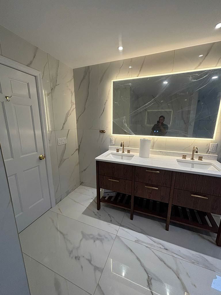 Modern bathroom with marble-style tile, double vanity, large backlit mirror, and a glass shower door.