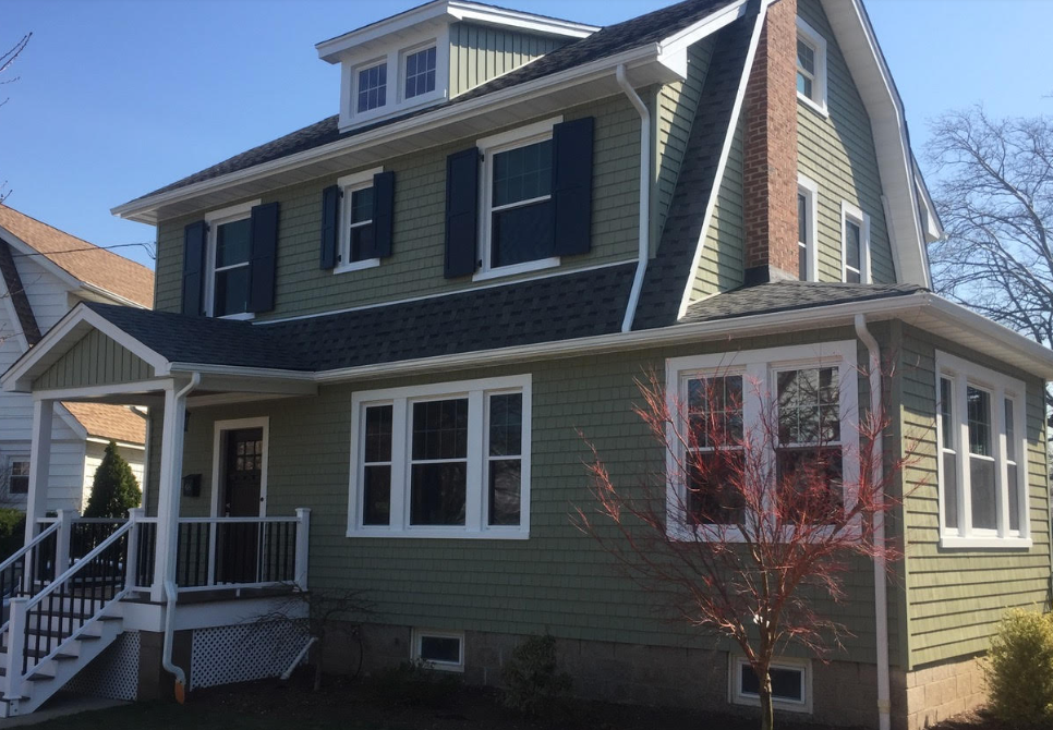 Green two-story house with white trim, front porch, and dormer windows in daylight
