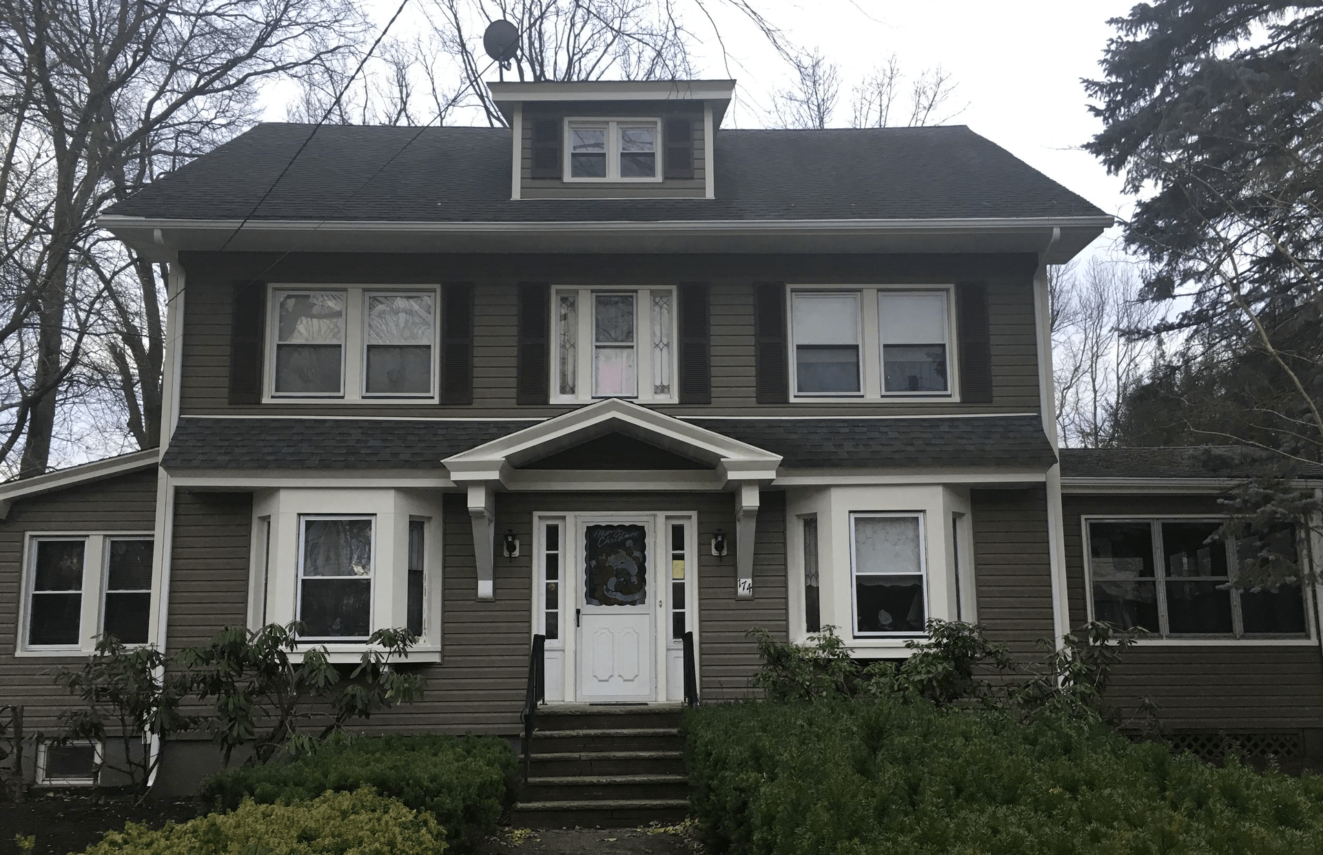 Large dark-gray Victorian-style house with white trim, front steps, and overgrown shrubs under bare trees.