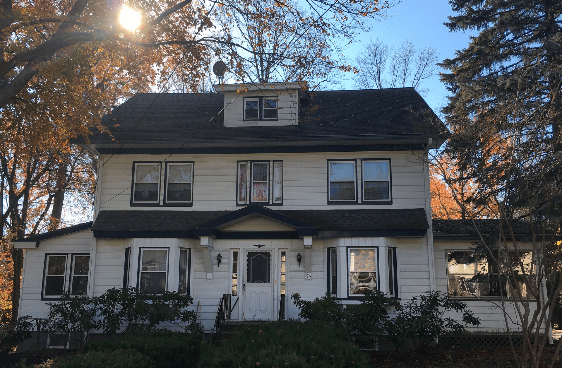 Two-story white house with dark roof and dormer, framed by trees in warm sunlight