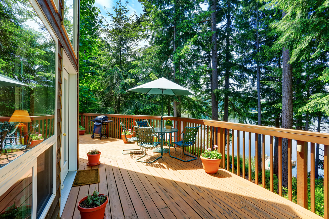 Wooden deck with outdoor table, green umbrella, potted plants, and forest view through railing