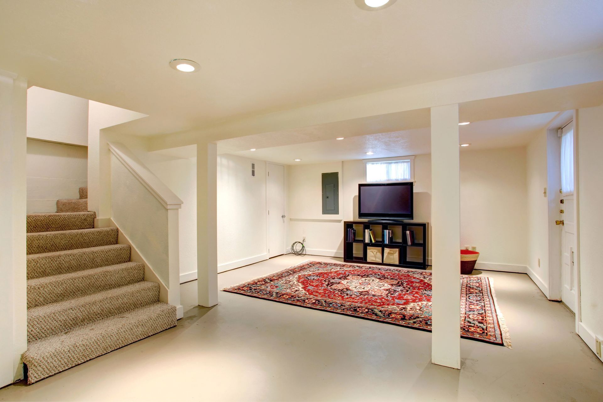 Finished basement rec room with beige carpeted stairs, white walls, and red patterned rug with TV area