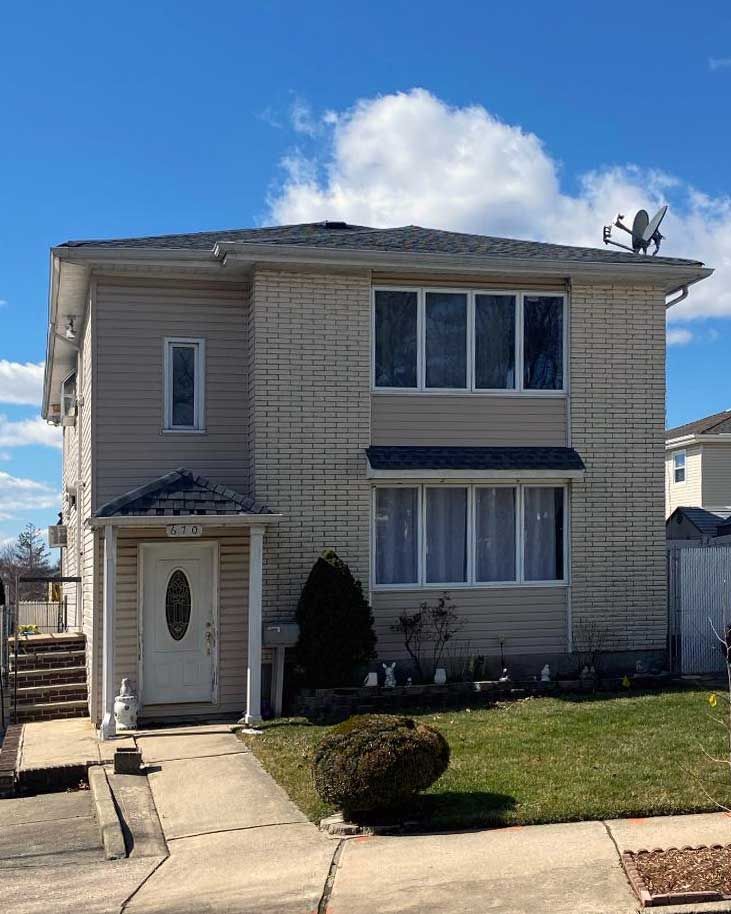 Two-story beige house with white trim, front door, windows, and a small lawn under a blue sky.