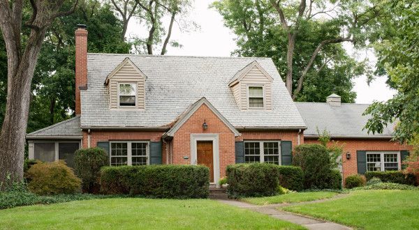 Brick house with gray roof, red front door, chimney, and trimmed hedges on a green lawn