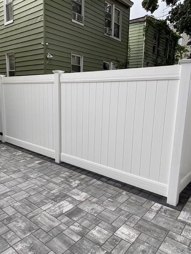 White vinyl fence beside a gray brick driveway in front of a green house