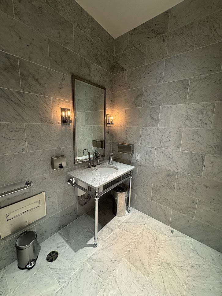 Modern bathroom with stone walls, wall mirror, sink, wall sconces, and a large marble-tiled floor.