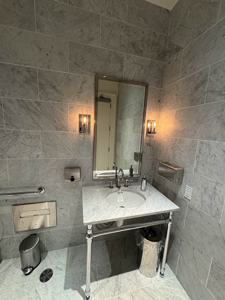 Bathroom sink with mirror, gray tile walls, wall sconces, and paper towel dispenser