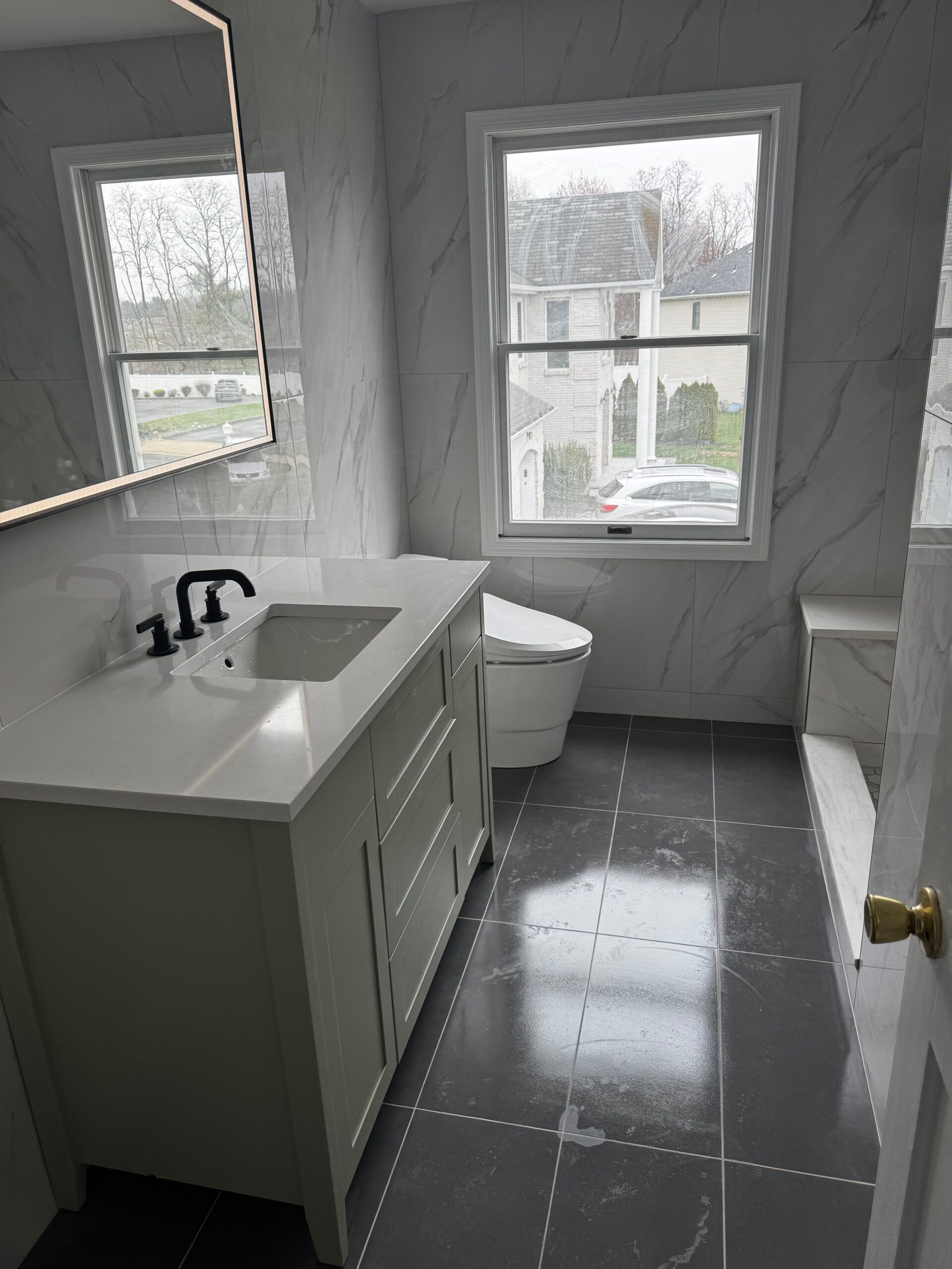 Small bathroom with white vanity, toilet, and window, featuring gray marble-patterned walls and dark tile floor