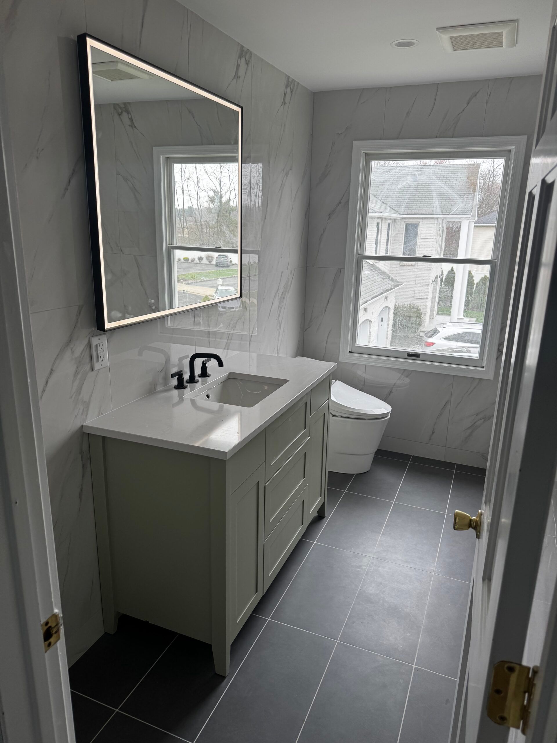 Small bathroom with green vanity, mirror, sink, toilet, and two windows over gray tile floor.