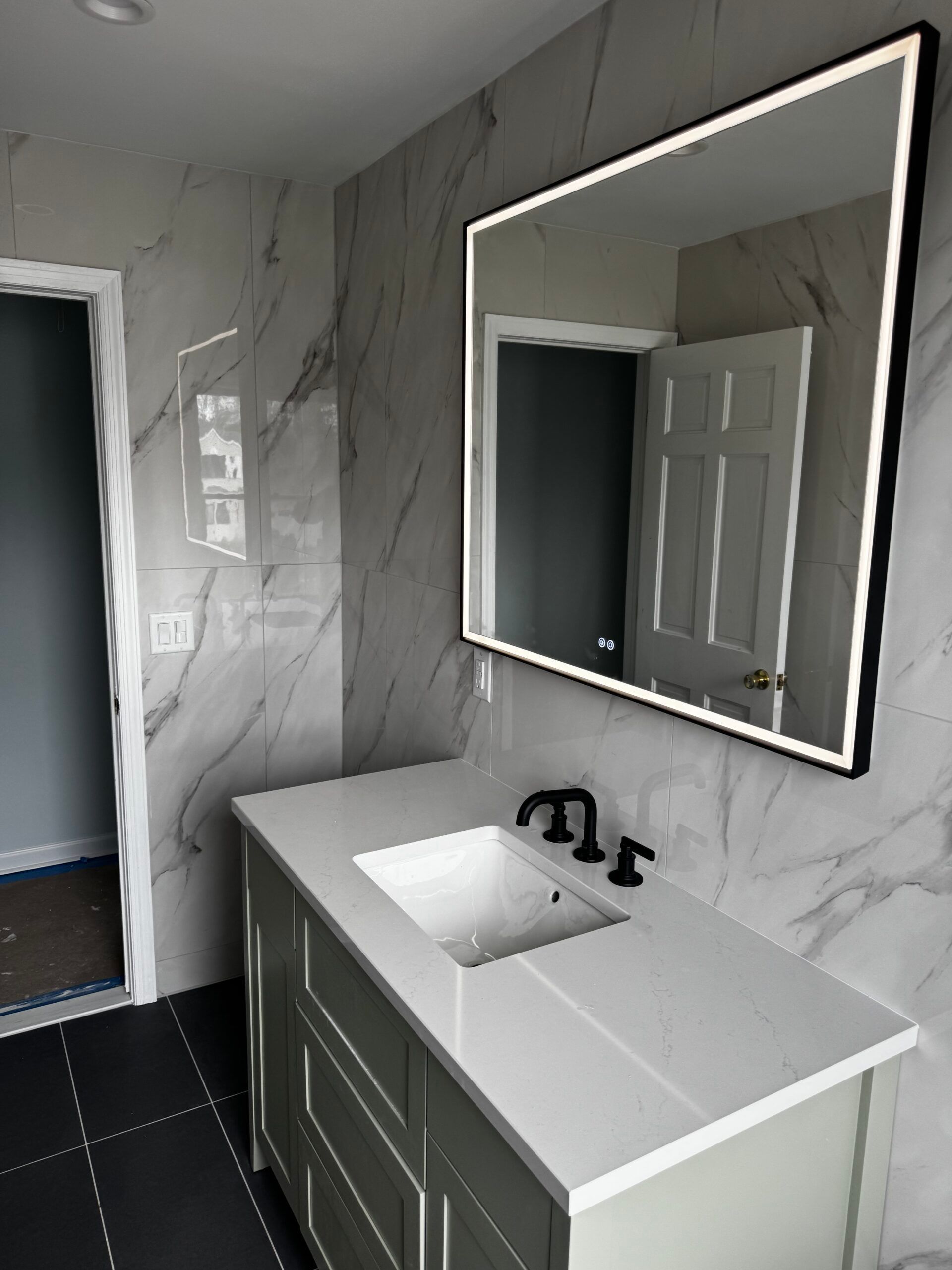 Modern bathroom vanity with gray marble walls, white sink, and large lit mirror