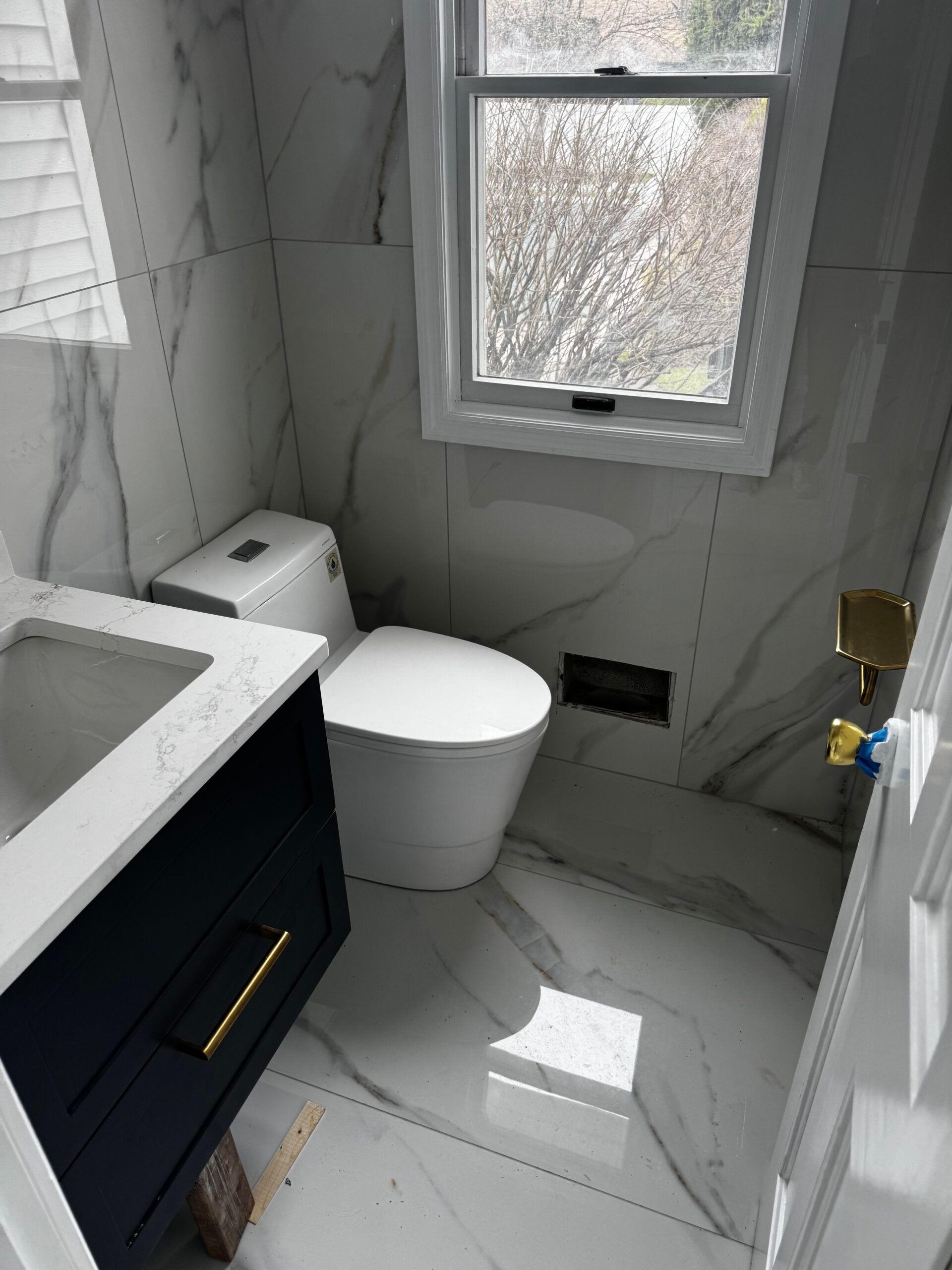 Compact modern bathroom with white tiled walls, black vanity, toilet, sink, and small frosted window.