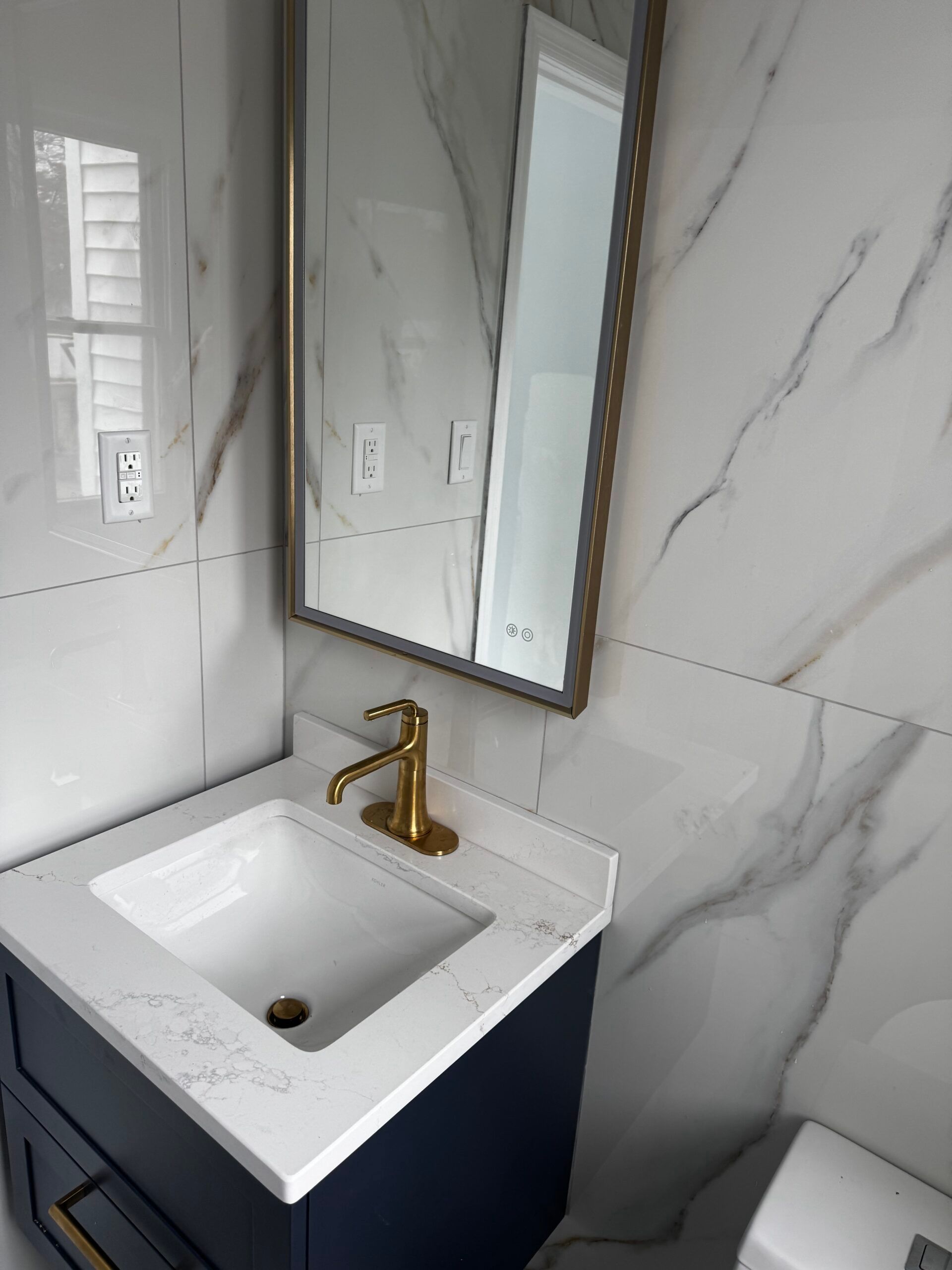 Small bathroom vanity with white sink, gold faucet, and wall mirror against marble-look tile walls