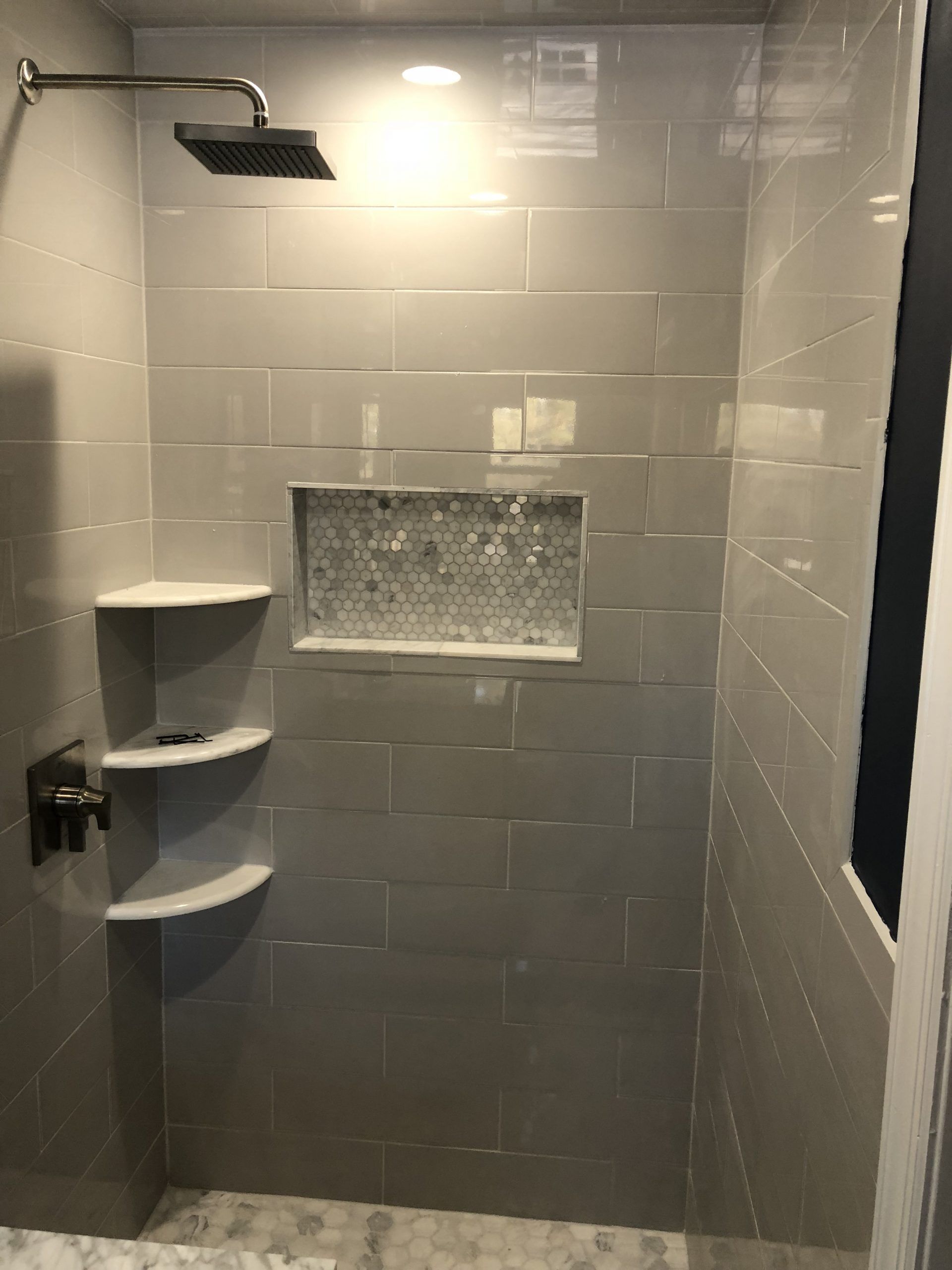 Gray-tiled shower with rain showerhead, built-in corner shelves, and recessed niche under bright light