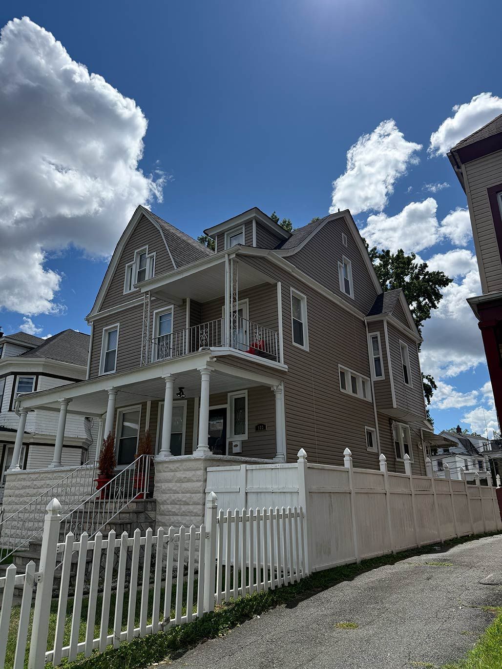 Victorian-style gray house with white trim, picket fence, and gravel driveway under a blue sky with clouds