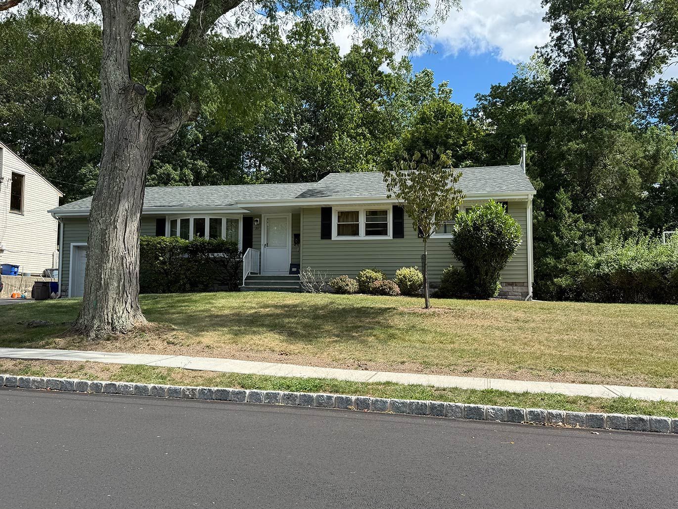 Single-story light green house with white trim, front porch, and large tree in the yard