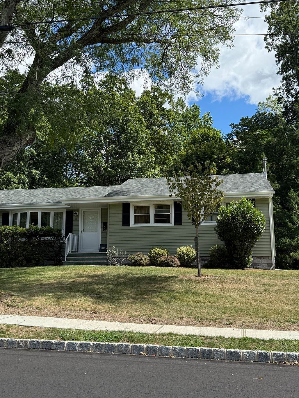 Single-story beige house with white trim, front porch, and large tree in a suburban yard