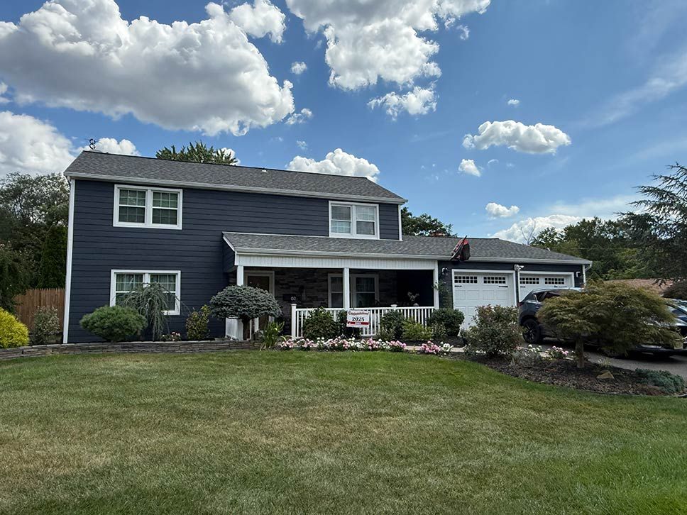 Two-story dark blue house with a white porch and garage, set behind a large grassy lawn under a cloudy sky