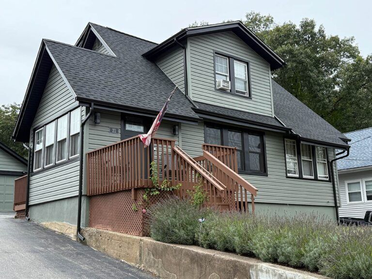 Gray house with steep roof, front porch, and wooden stairs beside a driveway.