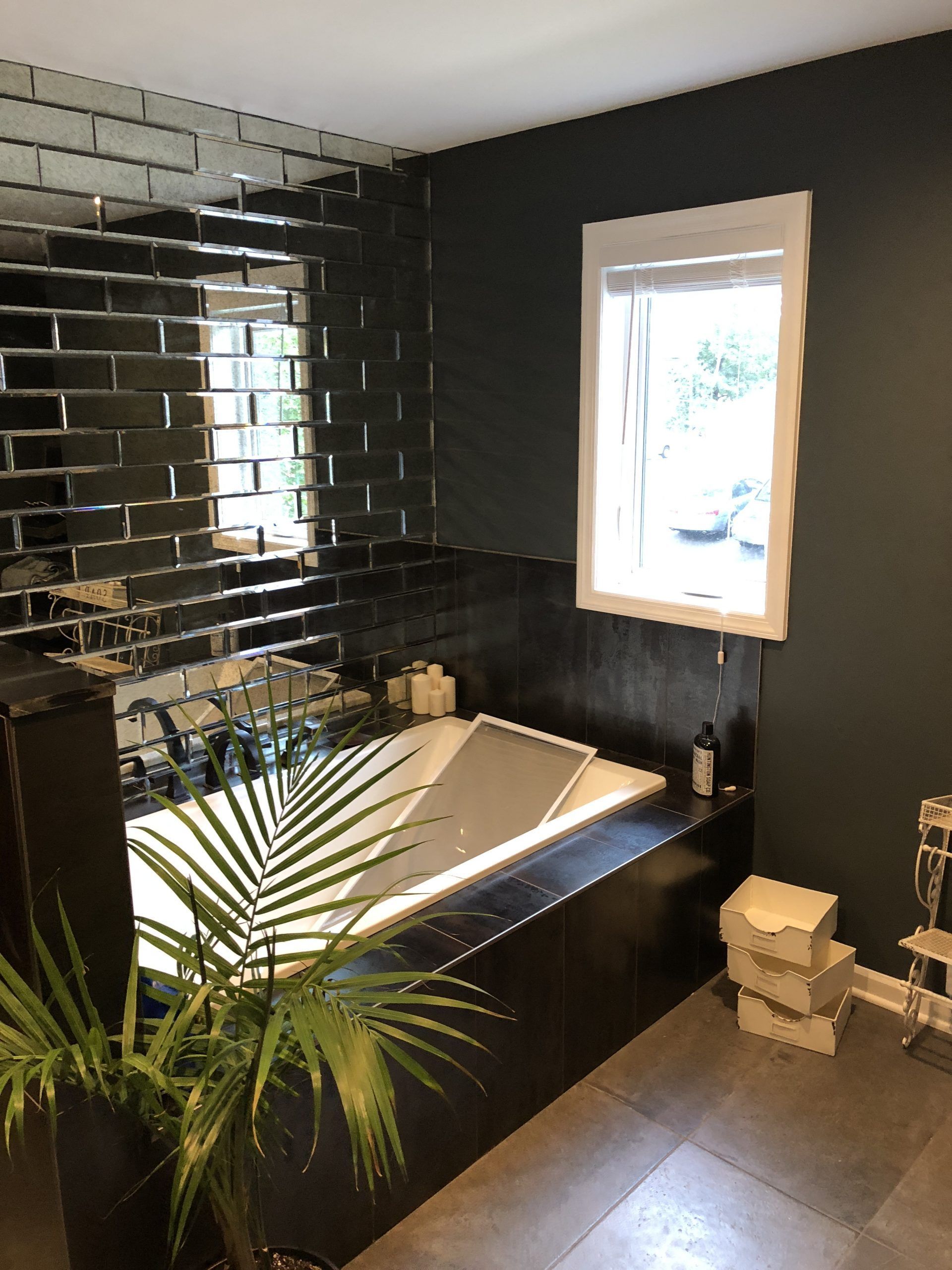 Modern black-tiled bathroom with a white bathtub, window, plant, and stacked towels
