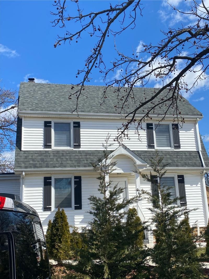 White two-story house with gray roof and shutters under a blue sky, framed by bare tree branches.
