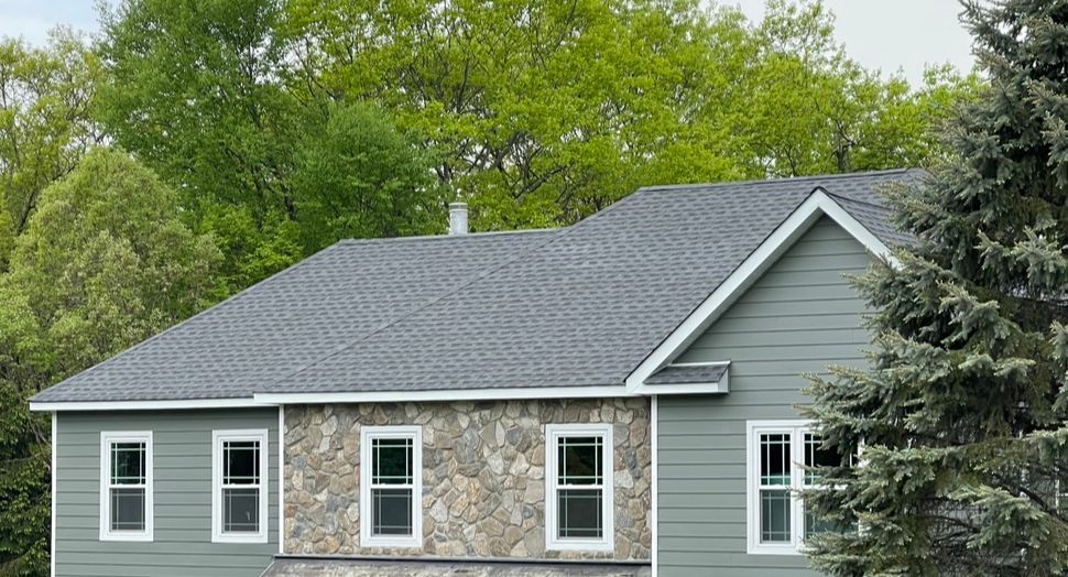 Gray house with stone facade and dark roof, surrounded by green trees and shrubs