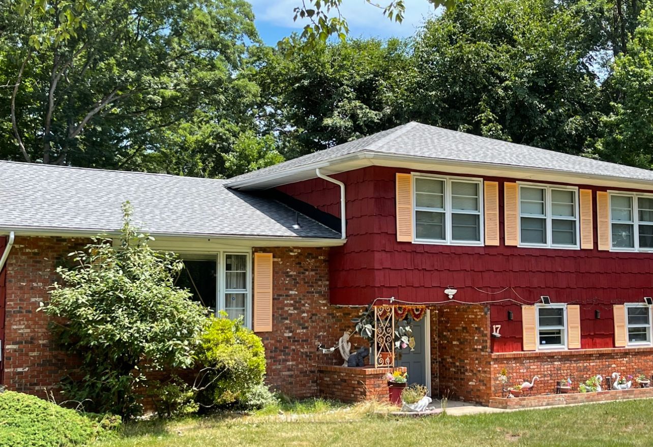 Suburban house with red siding, brick lower level, and a gray shingled roof surrounded by trees