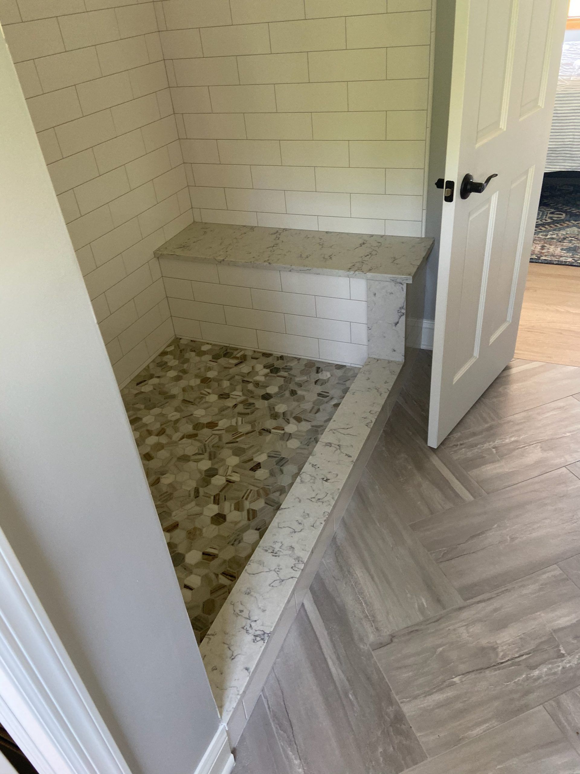 Small tiled shower with a bench and open white door in a bright bathroom.