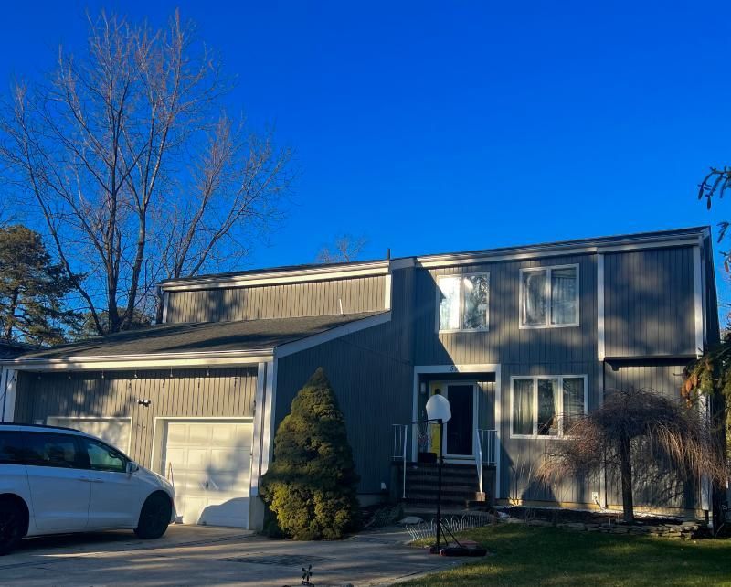 Two-story suburban house with gray siding, garage, driveway, and a white car under a clear blue sky