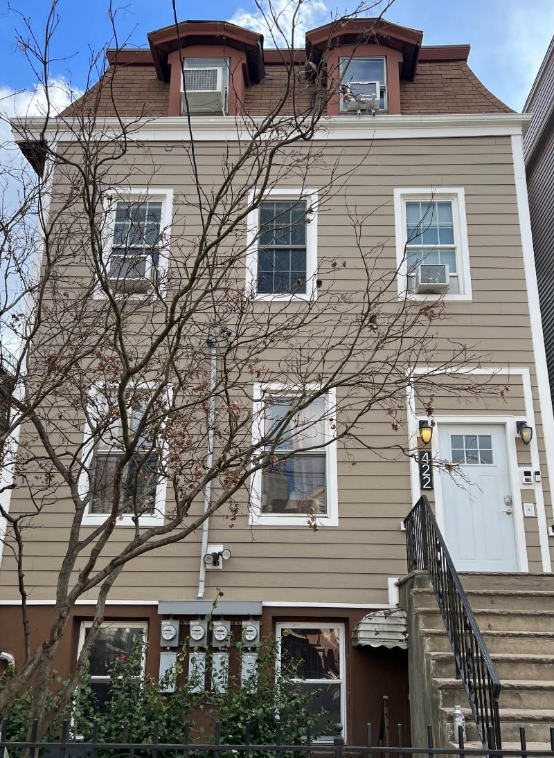 Three-story beige townhouse with white trim, stairs, leafless tree, and dormer windows under a cloudy sky