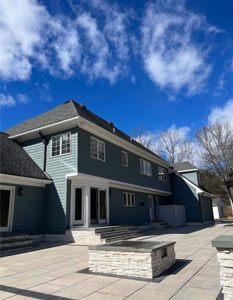 Blue two-story house with large patio and stone fire pit under a partly cloudy sky