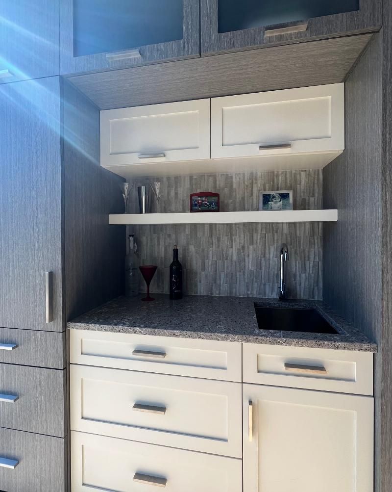 Modern kitchenette with white cabinets, gray tile backsplash, open shelf, sink, and granite countertop.