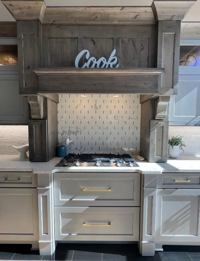 Kitchen stove beneath rustic wood range hood with “Cook” sign and tiled backsplash