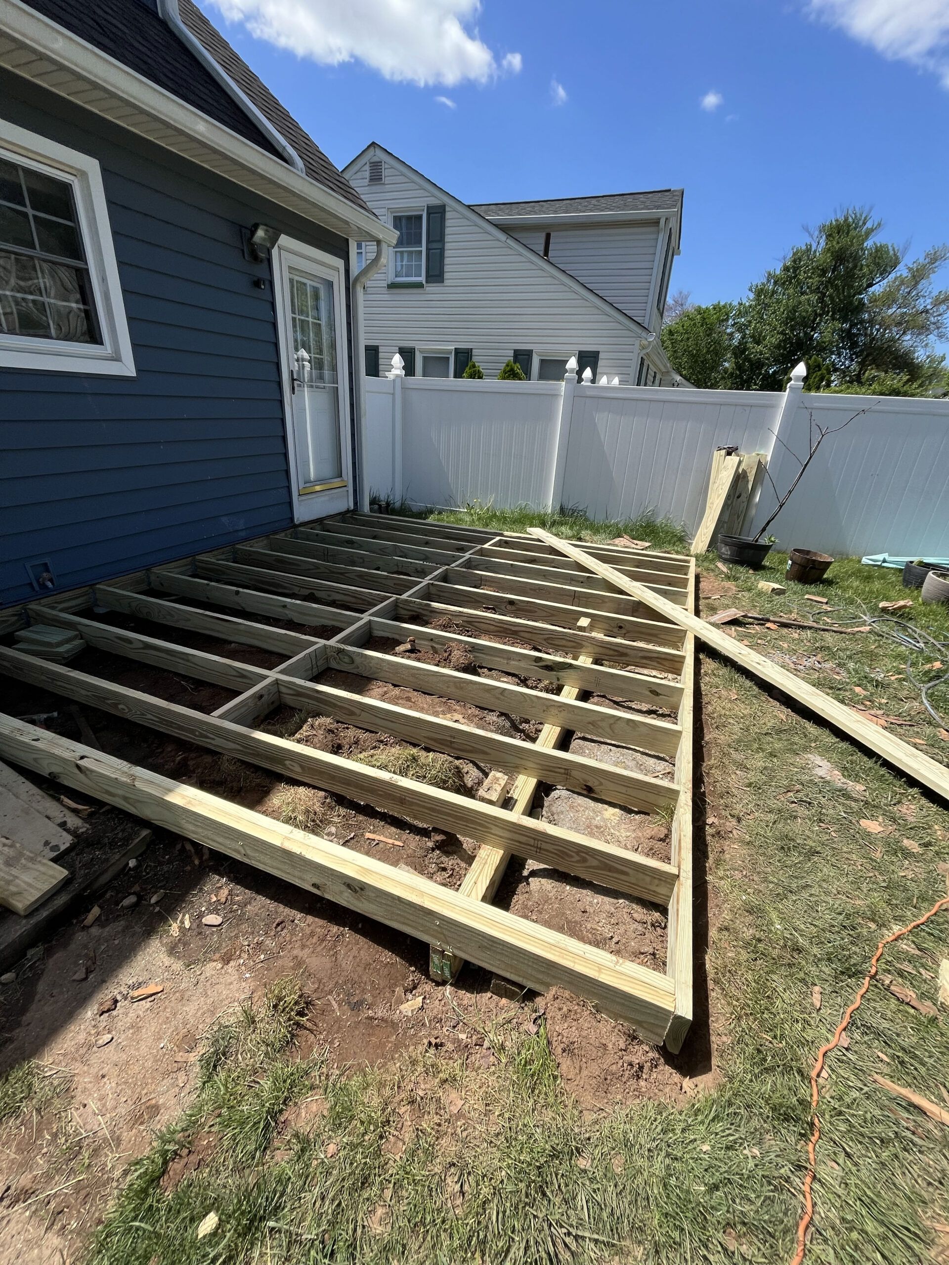 Backyard deck framing beside a blue house, with white fence and gravel ground