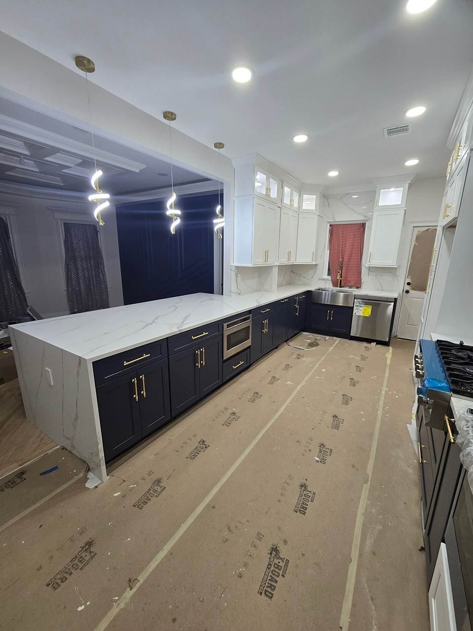 Empty kitchen with white ceiling lights, dark lower cabinets, white counters, and protective floor covering.