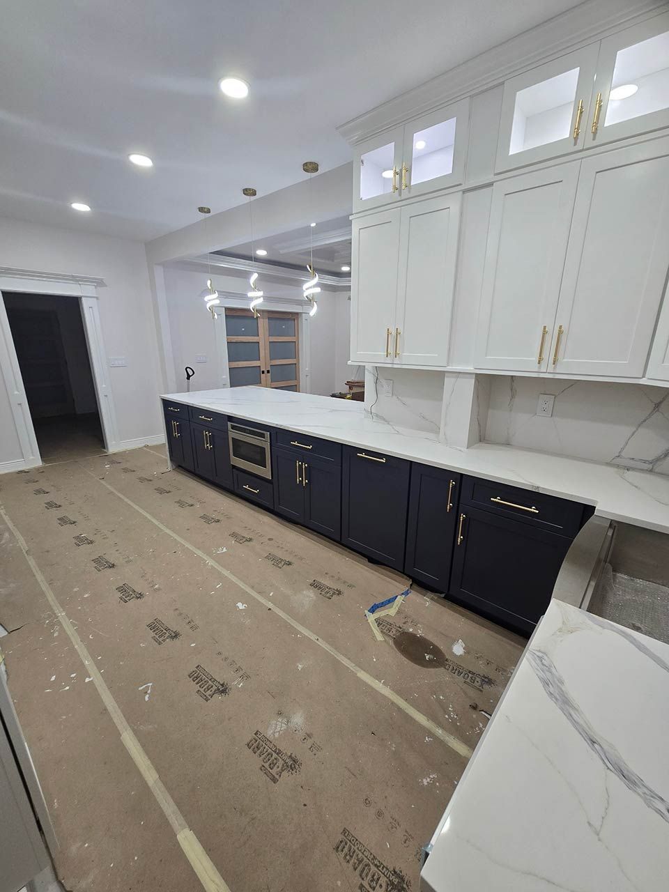 Kitchen under renovation with white upper cabinets, dark lower cabinets, and protective floor covering