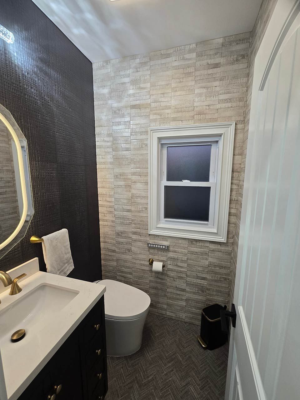 Small bathroom with stone-pattern walls, white vanity, toilet, and window under a sloped ceiling.