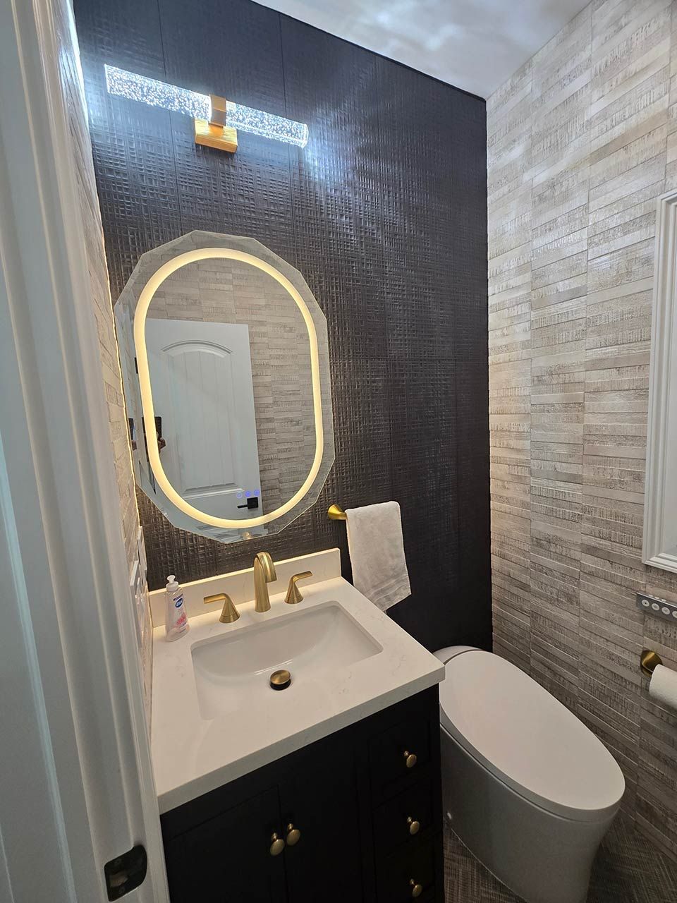 Small bathroom with dark mosaic accent wall, lit oval mirror, white vanity, and toilet beside shower tile.