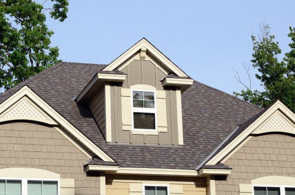 Suburban house roof with beige dormer window and dark gray shingles against blue sky
