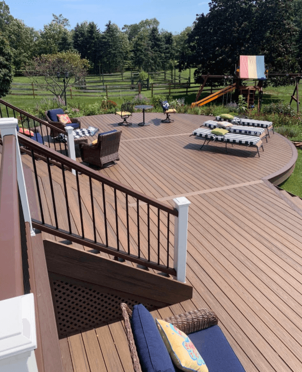 Wooden deck with patio furniture, picnic table, colorful play set, and grassy backyard trees.