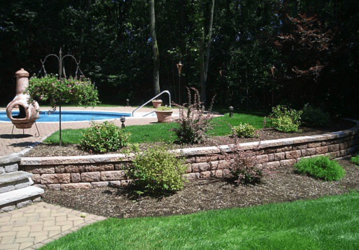 Backyard pool with stone retaining wall, shrubs, and green lawn surrounded by trees