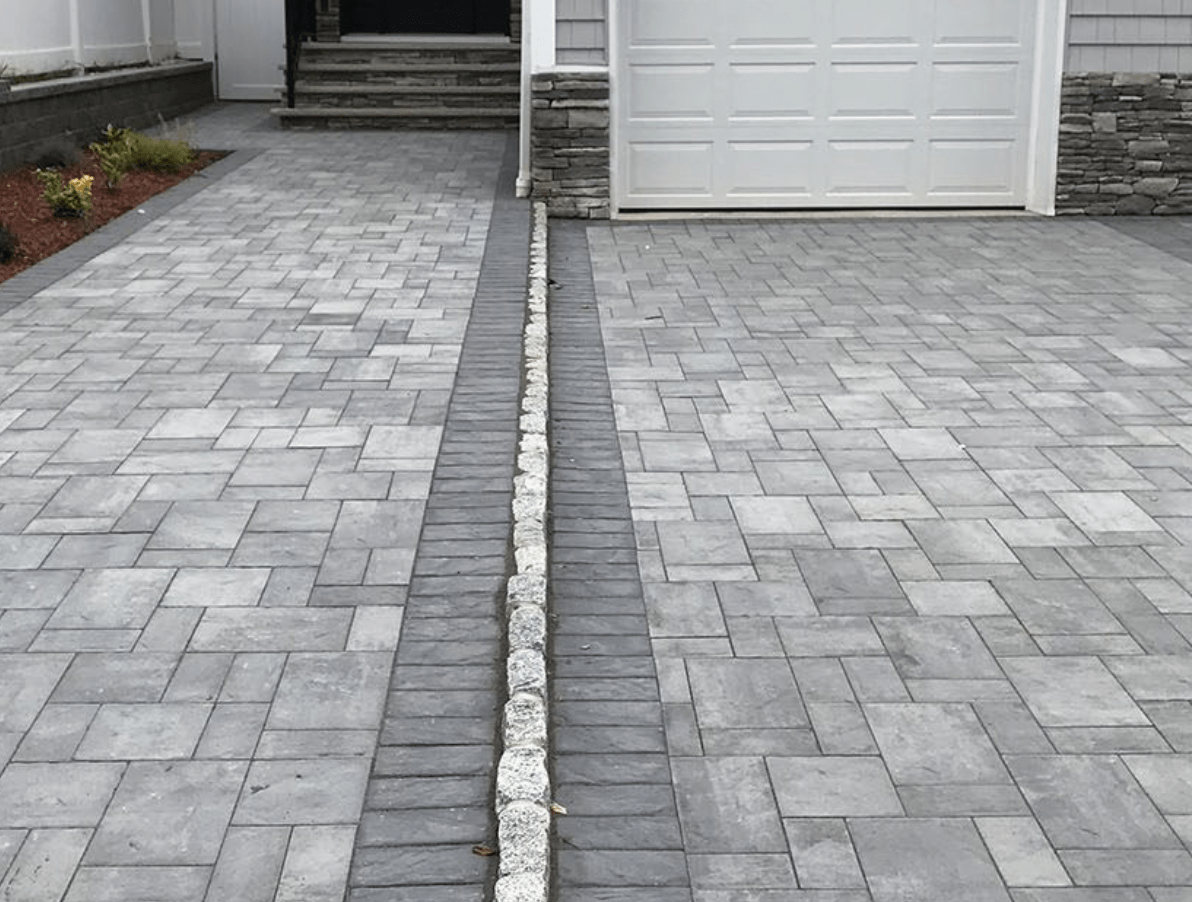 Gray paver driveway split by a narrow drainage trench leading to a white garage door