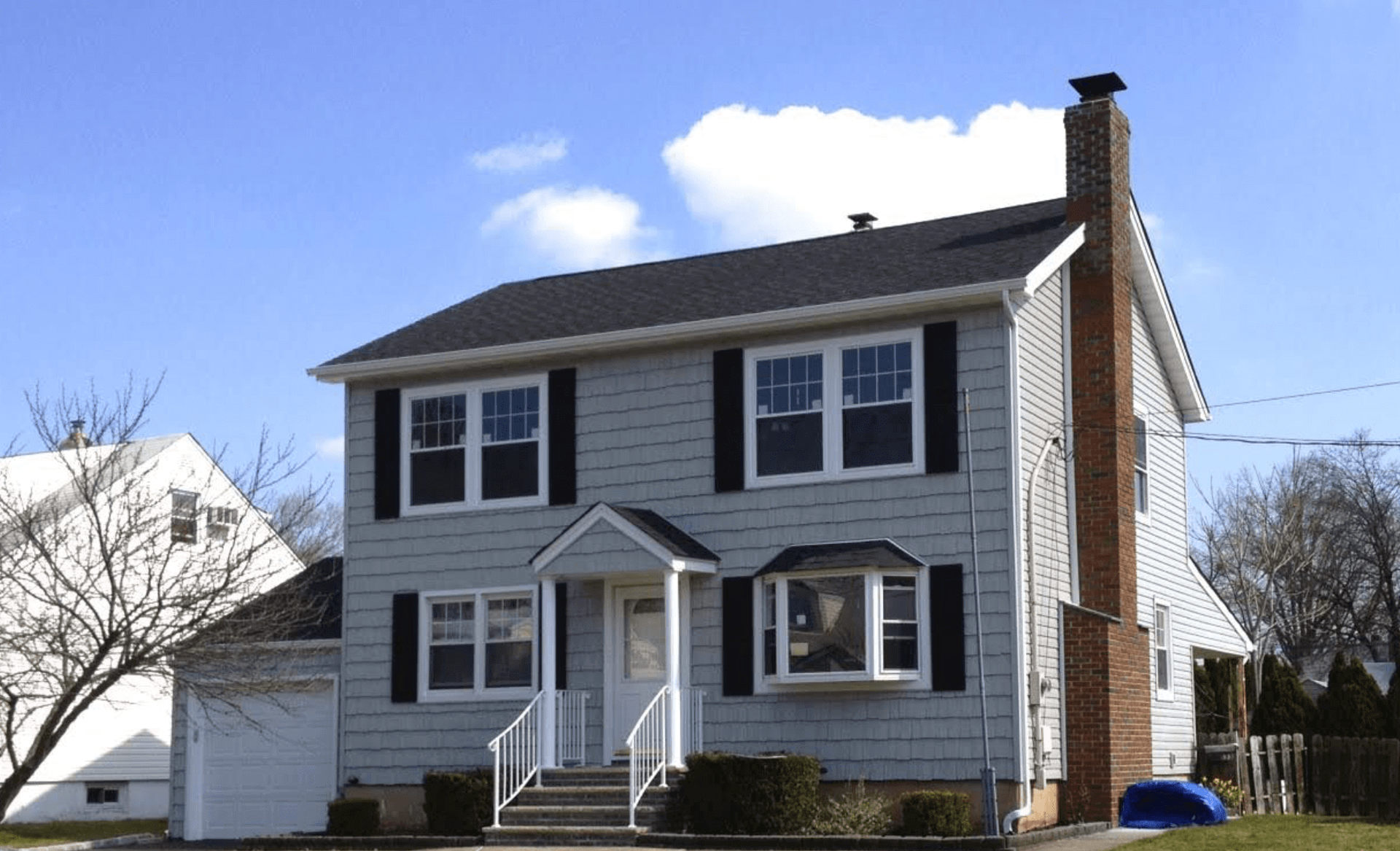 Two-story white house with black shutters, front porch, garage, and chimney under a blue sky