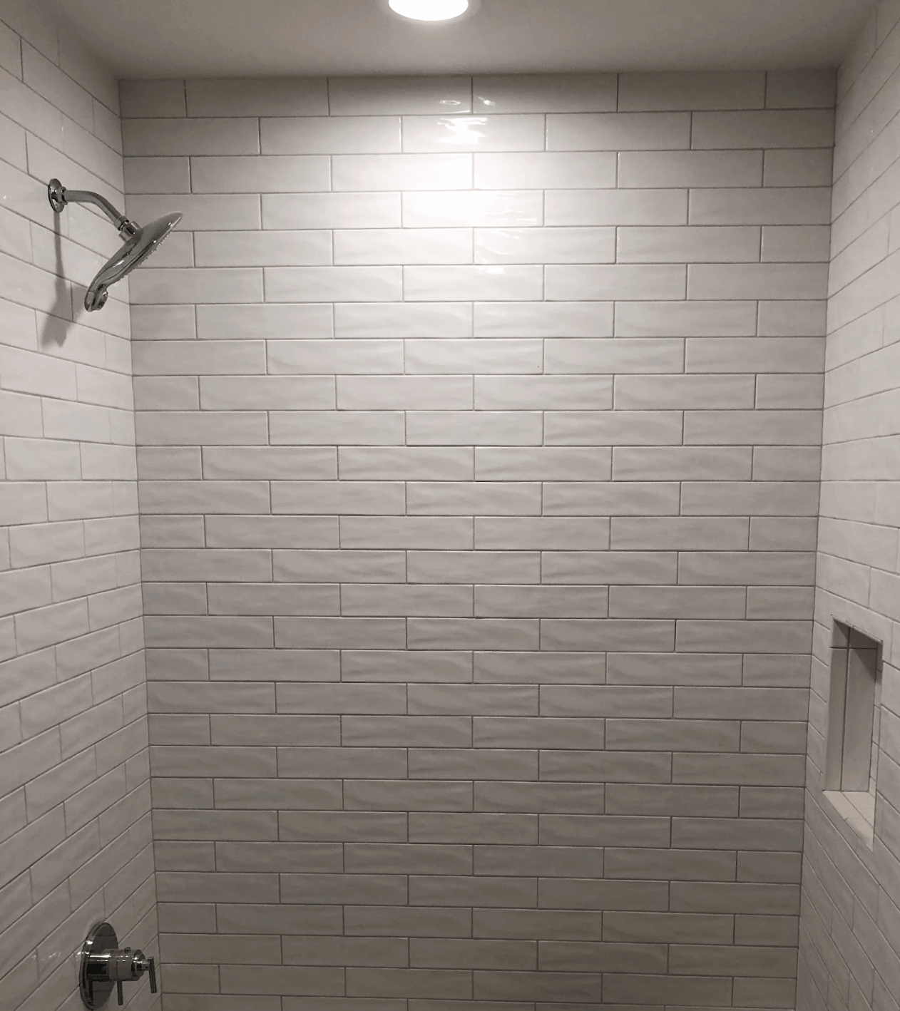 Empty tiled shower with a wall-mounted showerhead and recessed shelf, lit by a ceiling light.