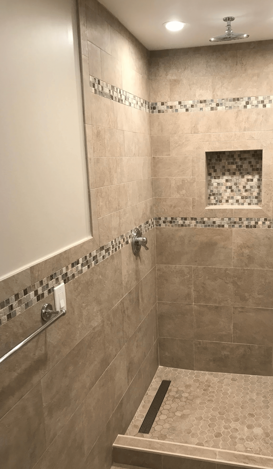 Bathroom with beige tiled shower, recessed shelf, and decorative border tile under ceiling lights
