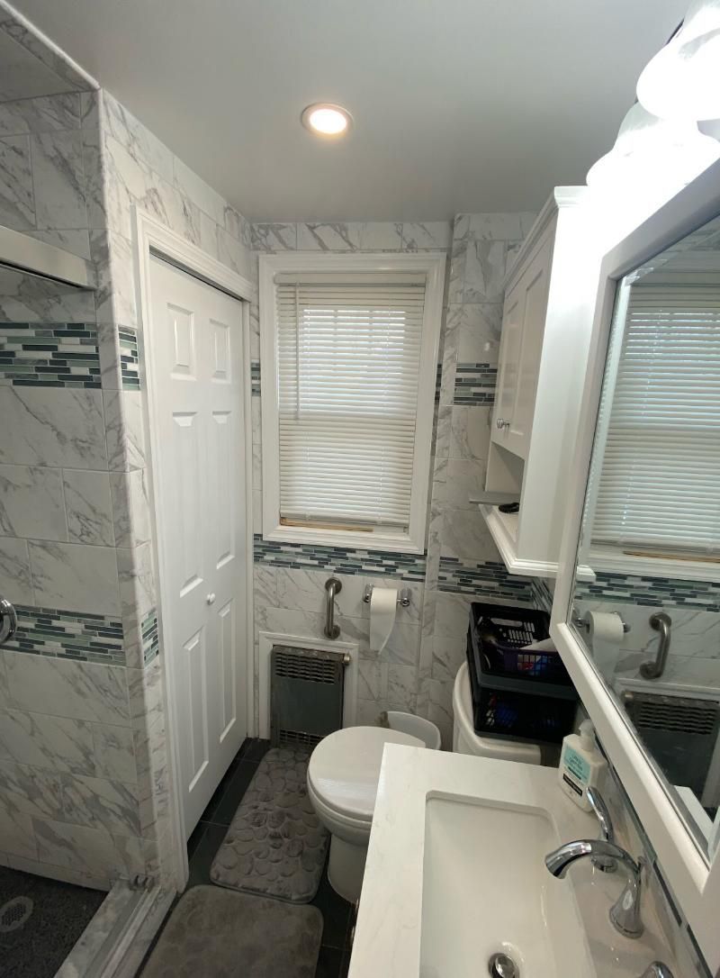 Narrow white bathroom with marble-pattern walls, window blinds, toilet, sink, and vanity mirror.