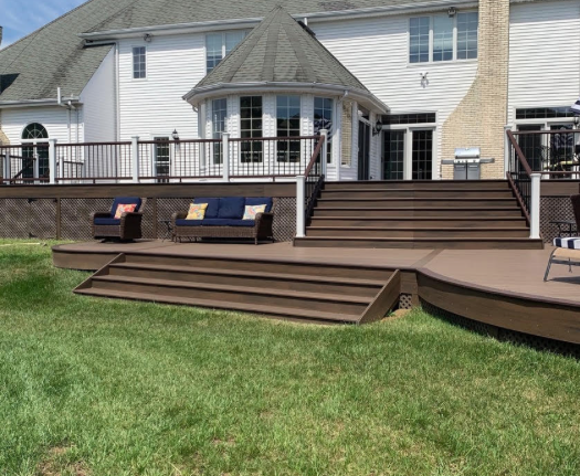 Backyard deck with multiple brown stairs and white house siding, bordered by green lawn and patio seating