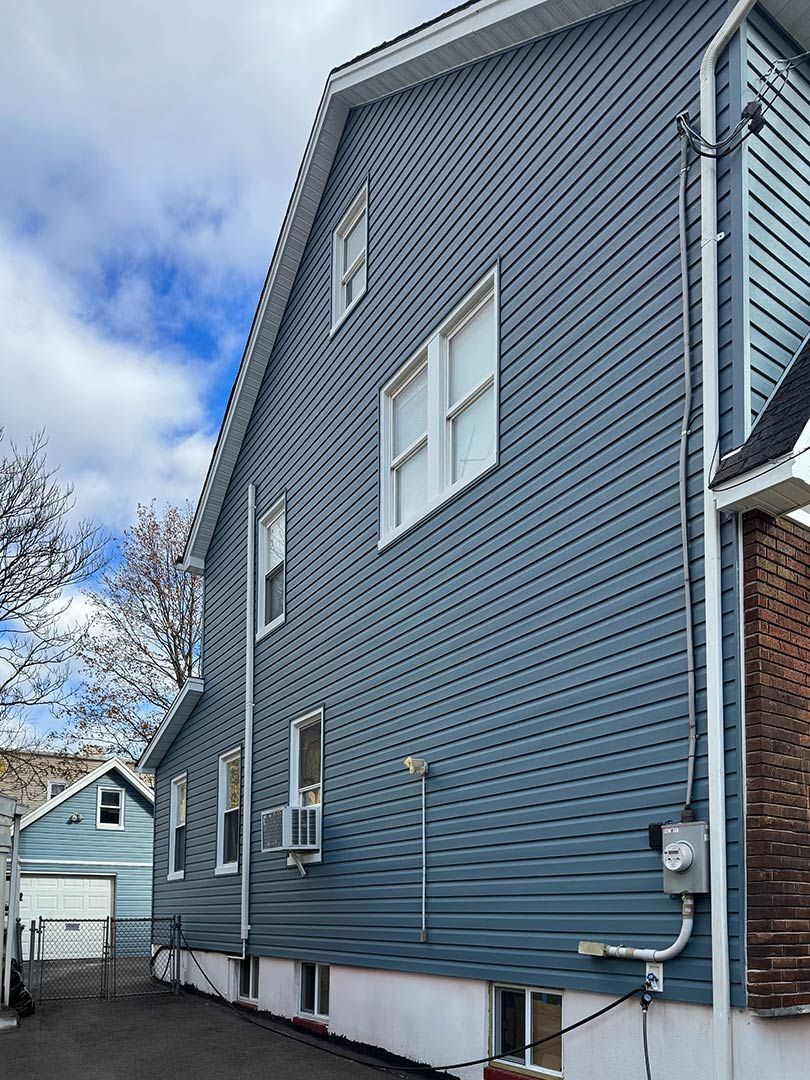 Blue-sided house exterior with white-trim windows, air conditioners, and a narrow side yard under a cloudy sky