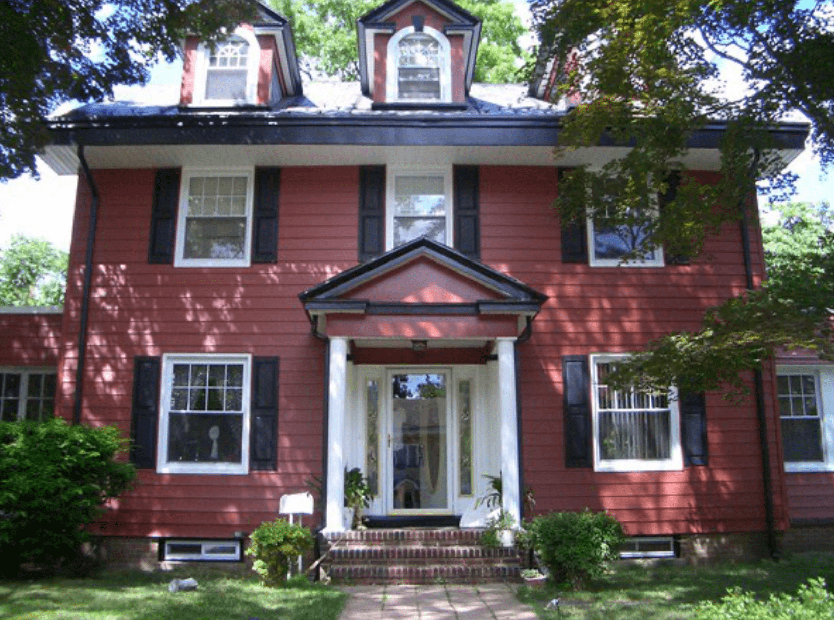 Red two-story house with white trim, dormer windows, and a small front porch surrounded by trees.