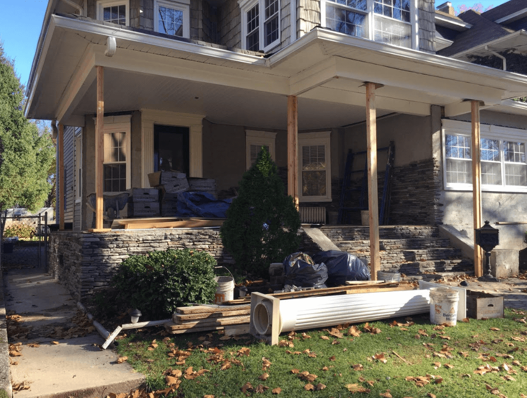 Front porch of a house with stone steps, columns, and outdoor seating, surrounded by autumn leaves.